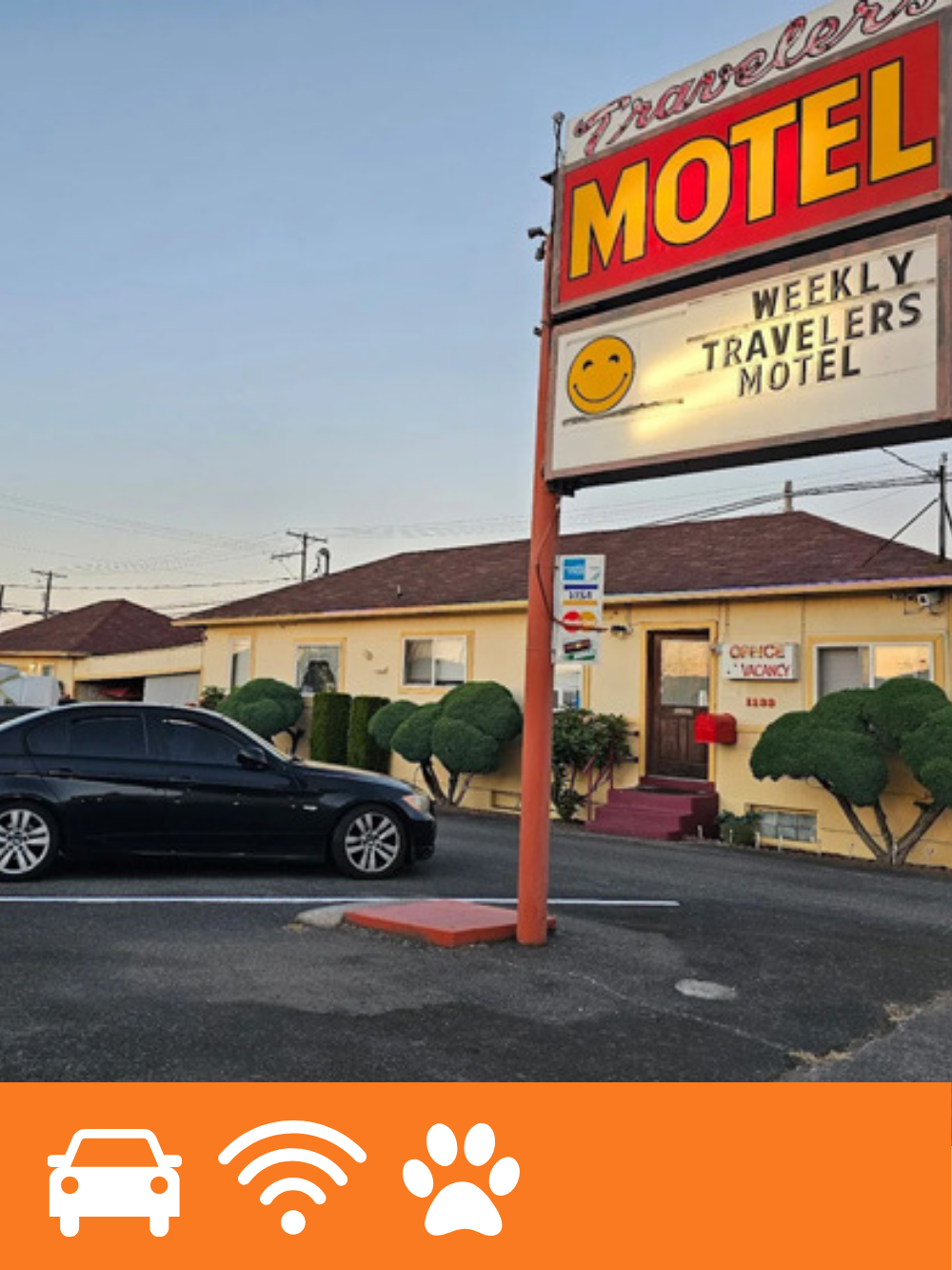 A vintage roadside motel sign advertising Travelers Motel with a smiley face, positioned in front of a yellow single-story building with landscaping and a parking lot with a black car.
