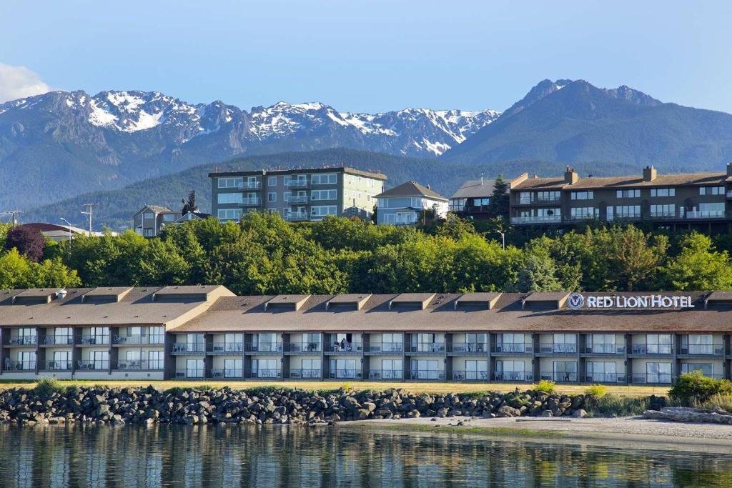 A hotel named Red Lion Hotel beside a body of water with rocky shoreline, green trees, and residential buildings, with snow-capped mountains in the background.