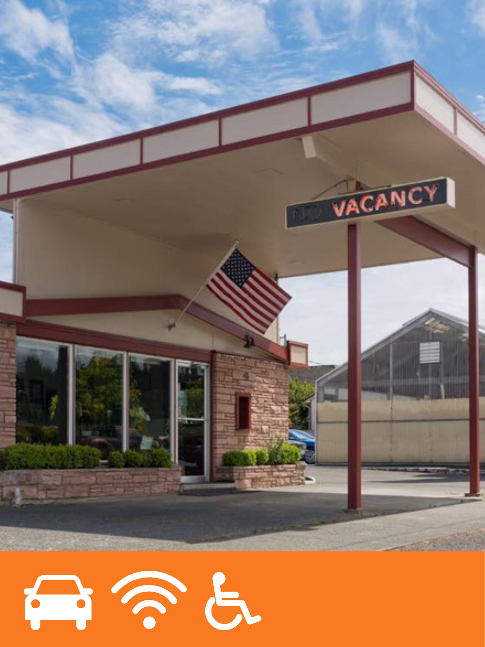 A small building with a brick facade, large windows, and a green hedge in front, with a canopy extending over the sidewalk. An American flag is mounted on the building, and a sign reads 'NO VACANCY'.