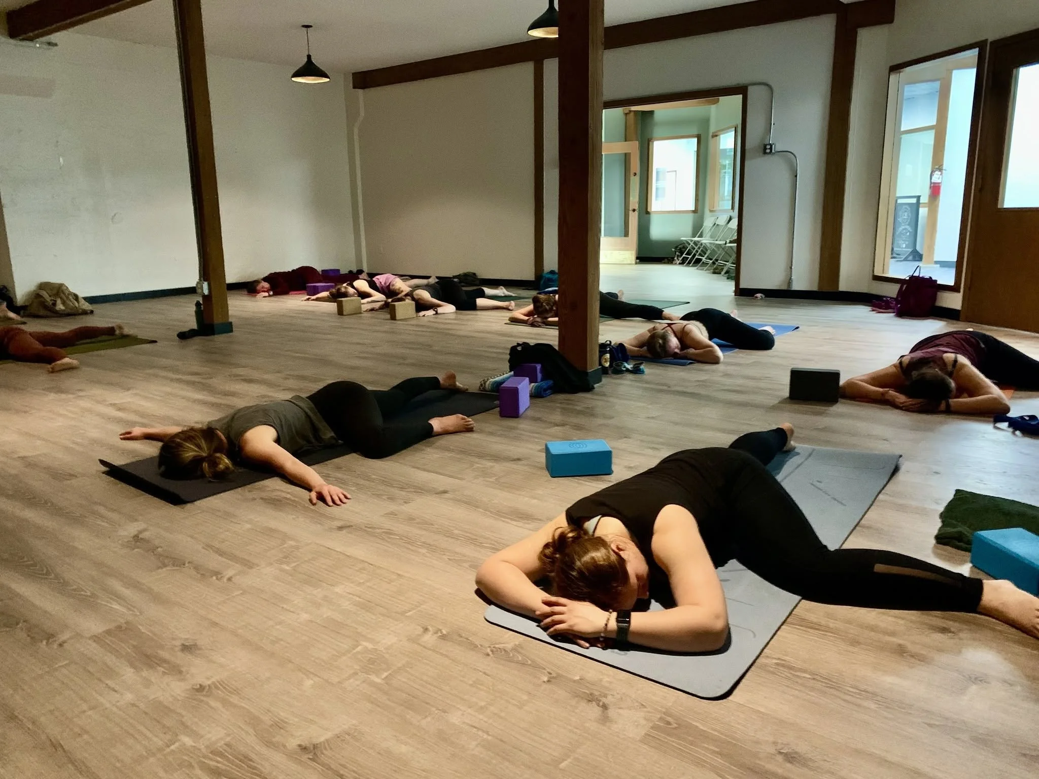 People participating in a yoga class outdoors on a wooden patio surrounded by plants and a colorful house with a tree in the background.