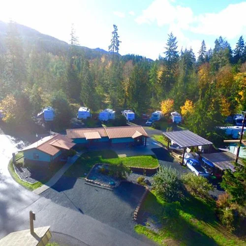 Outdoor view of a wooded area with parking spaces, camper trailers, and a building with a brown roof surrounded by trees and mountains in the background.
