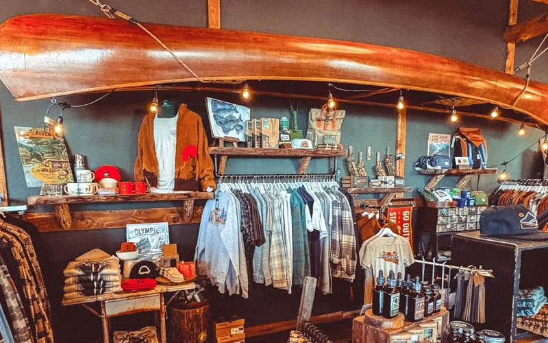 Interior of a retail store with clothing, accessories, and souvenirs, featuring a wooden canoe mounted as decor and warm string lighting.