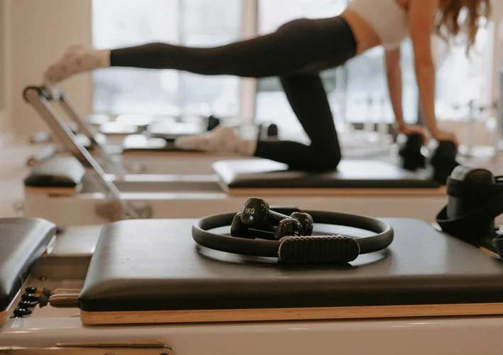 Close-up of a black fitness ring and dumbbell on a workout platform in a gym, with a woman exercising on a reformer machine in the background.
