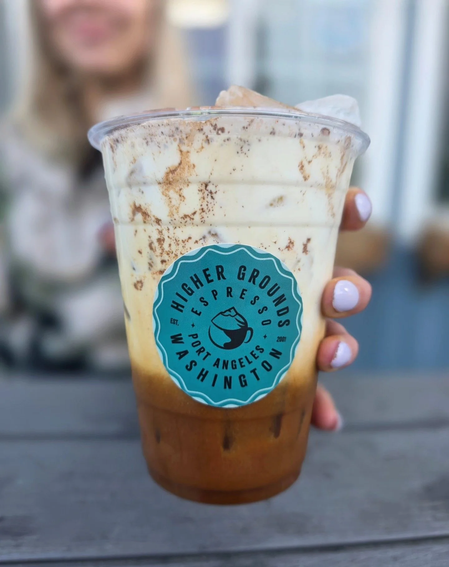 A large iced coffee or espresso drink in a to-go cup with whipped cream and caramel drizzle, held outdoors near a wooden building.