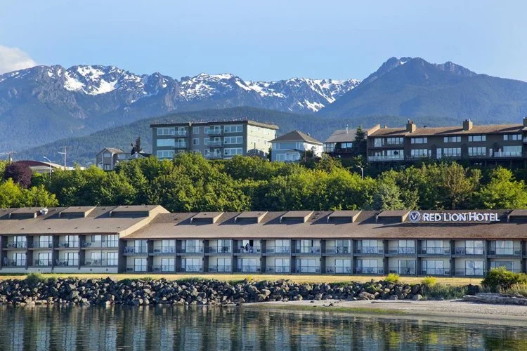 A hotel named Red Lion Hotel with multiple balconies along a waterfront, surrounded by green trees and set against snow-capped mountains under a blue sky.