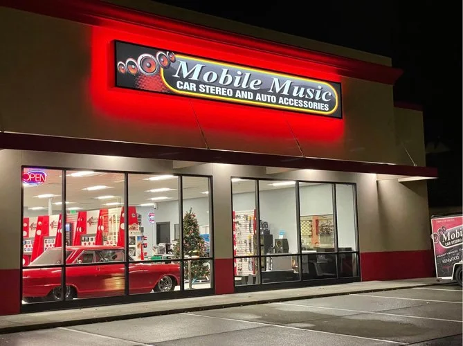 Exterior view of Mobile Music store at night, illuminated sign, large windows displaying a red vintage car inside, Christmas tree, and music accessories, with a red and white color theme.
