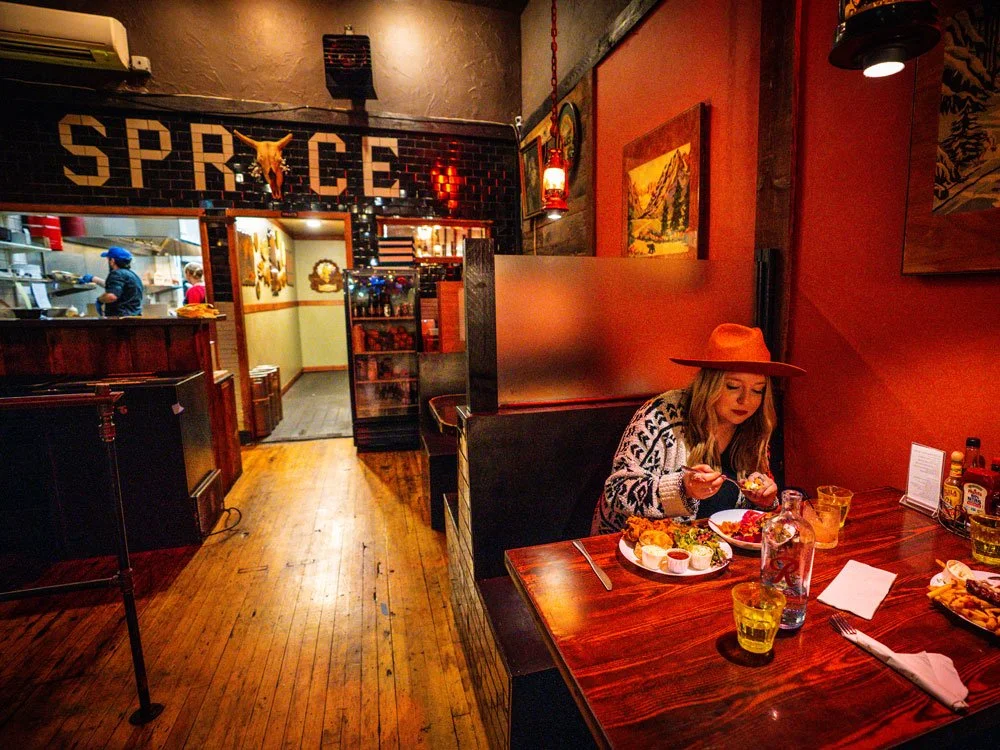 A woman with a large orange hat eating a meal at a restaurant with wooden tables and warm lighting.