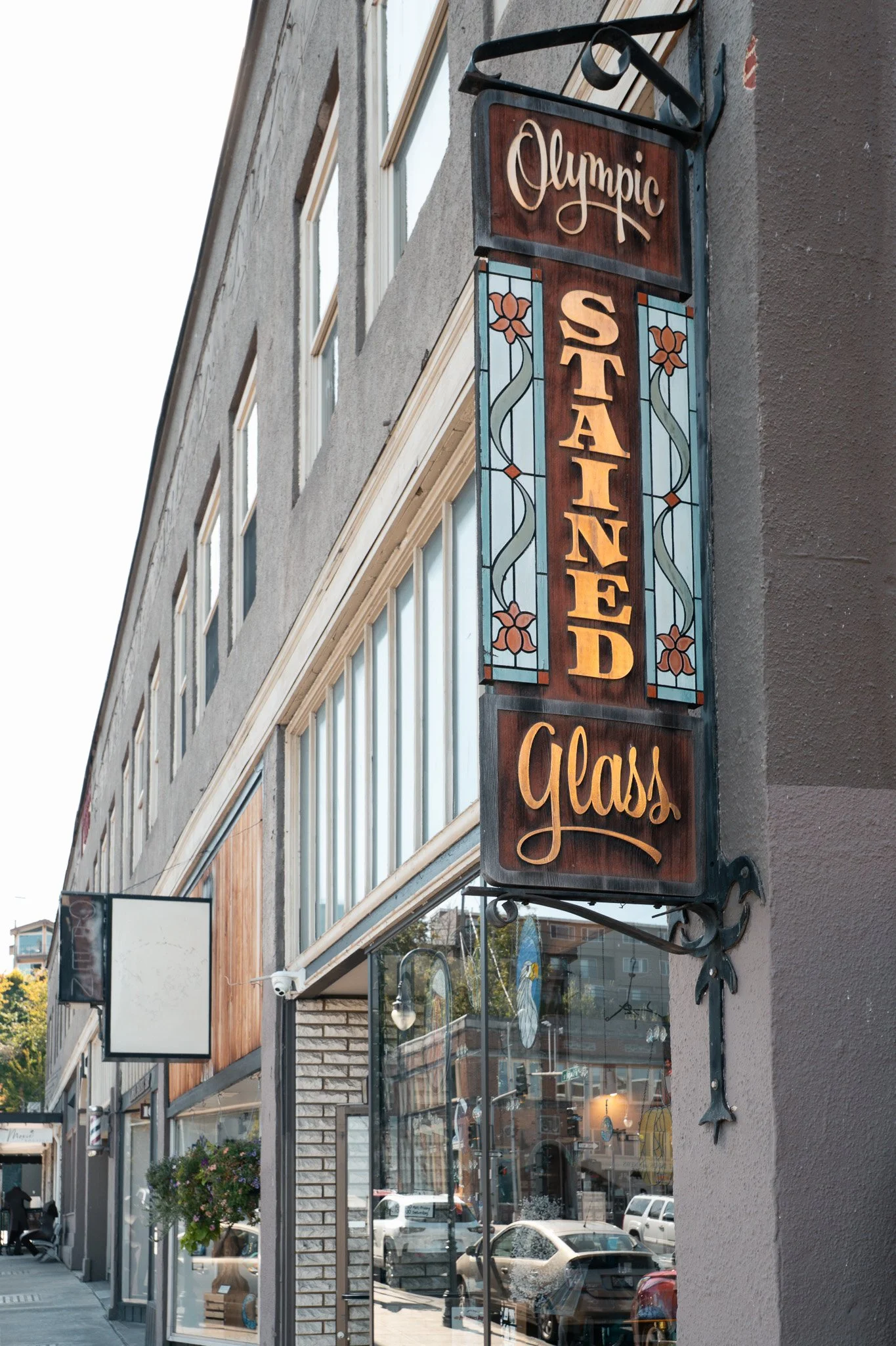 Street view showing storefronts on a sunny day, including a sign for Olympic Standing Glass and Harbor Art, with pedestrians walking.