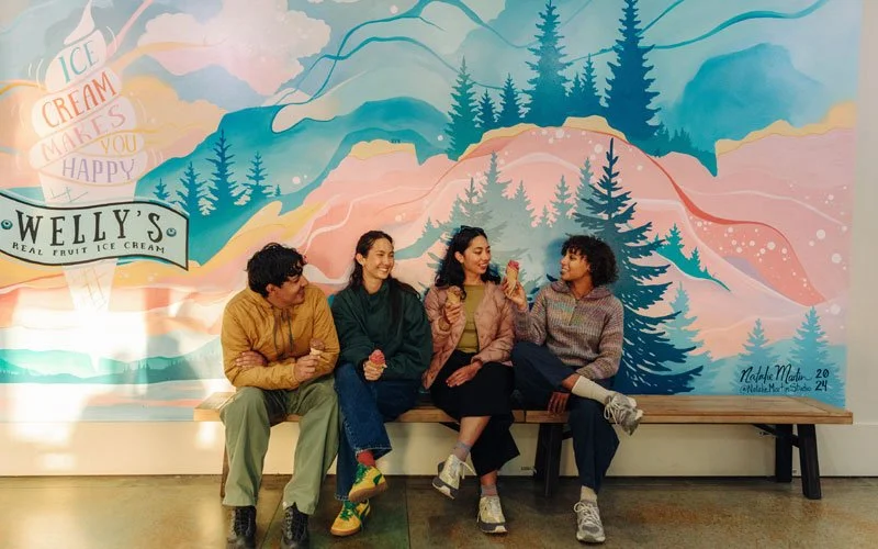 Four people sitting on a wooden bench, enjoying ice cream cones in front of a colorful mountain mural and trees.