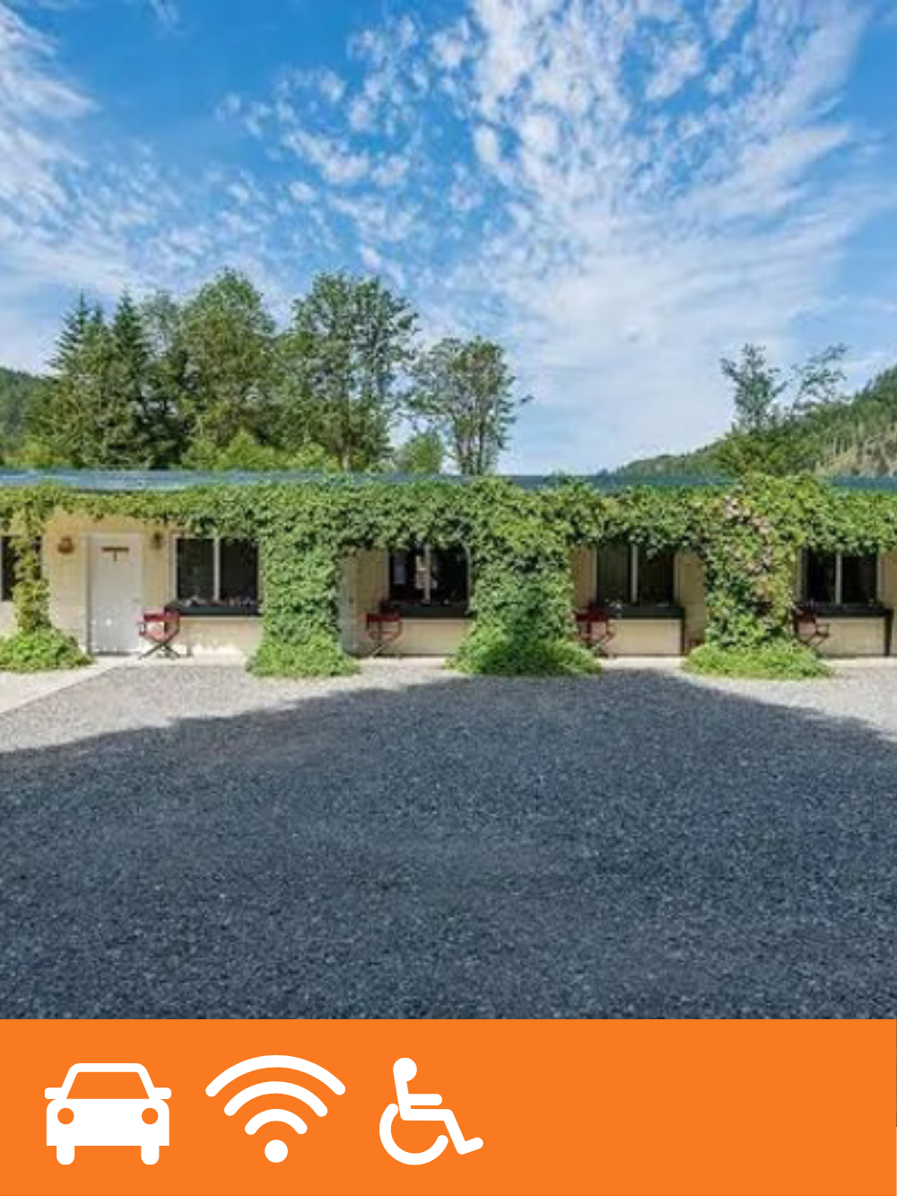 A building with a vine-covered porch and multiple windows, set in a natural landscape with trees and a blue sky with clouds.
