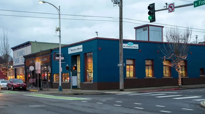 Street corner with blue building houses Waters West and Fly Fishing Shop, trees with string lights, cars parked, traffic lights, and street signs, in an urban area.