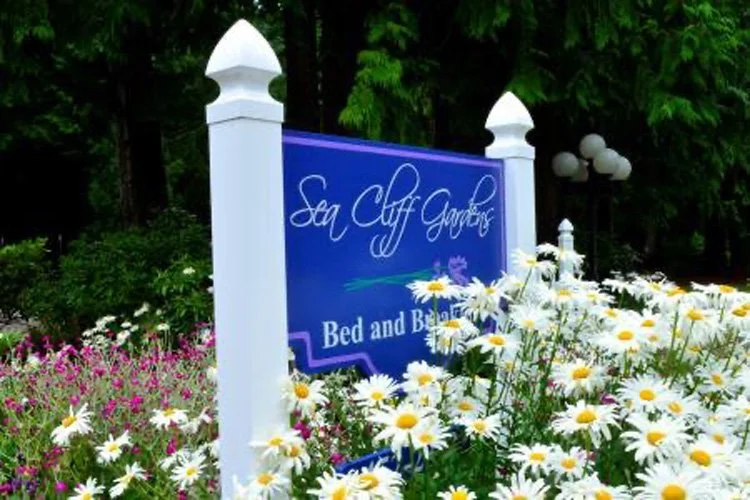 A Sign that reads 'Sea Cliff Gardens Bed and Breakfast' surrounded by white daisies and pink flowers, with green trees in the background.