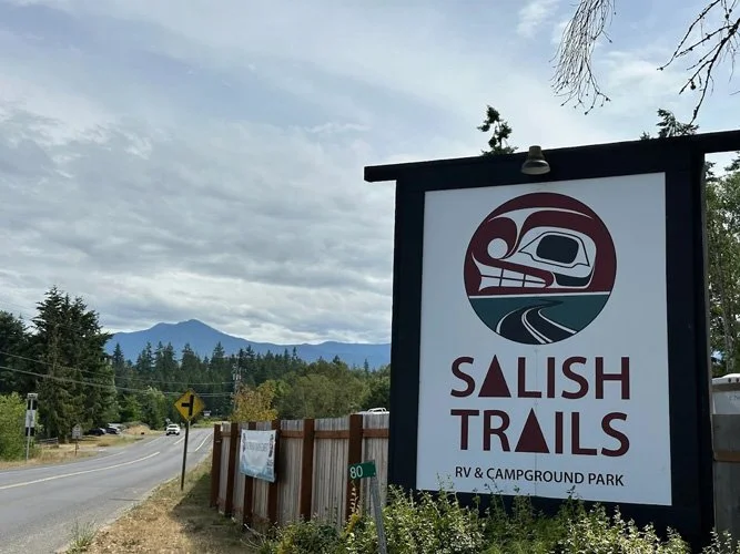 Sign for Salish Trails RV and campground park, featuring a stylized logo with a train and a mountain landscape, beside a country road with trees and distant mountains under cloudy skies.