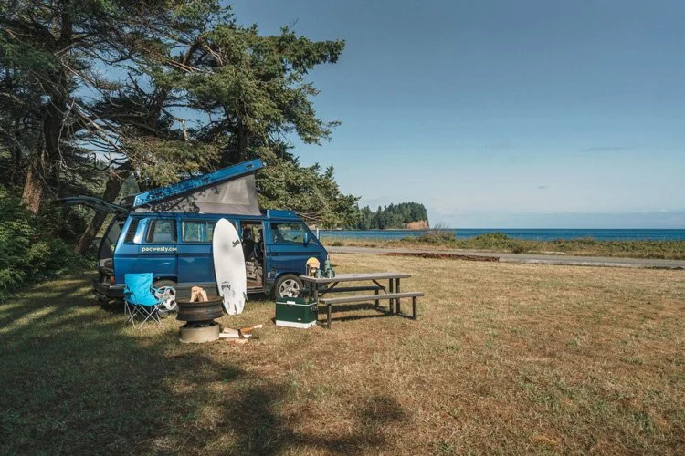 A camping site near the ocean with a blue van parked on a grassy area, an open hatch, a camping chair, a surfboard, and a picnic table.