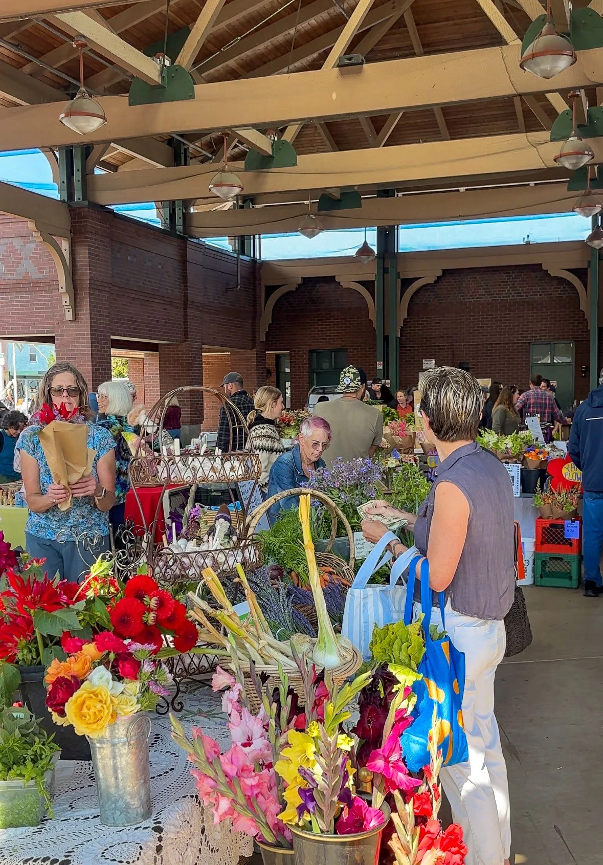 A lively scene under the Gateway Pavillion during the Port Angeles Farmers Market, with customers checking out farm stands with beautiful flowers and vegetables.