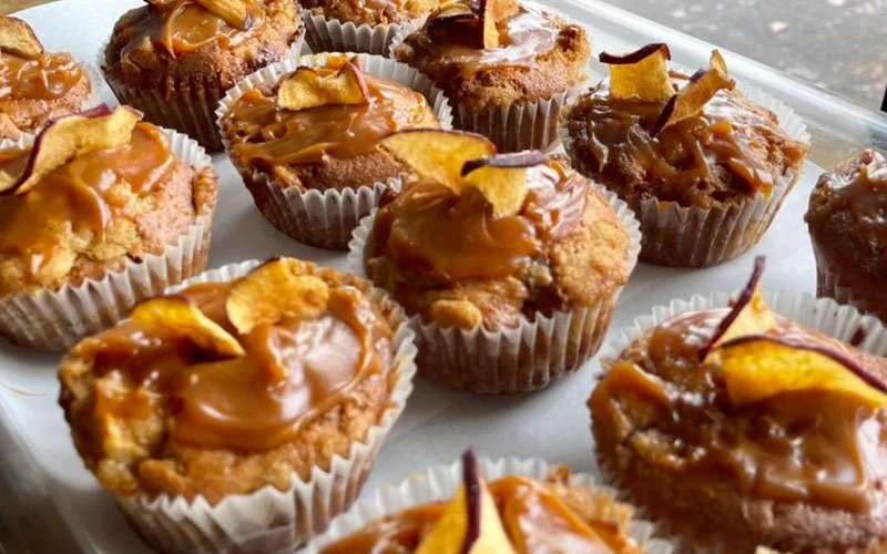 A tray of caramel apple cupcakes topped with caramel frosting and apple pieces, in white cupcake liners.