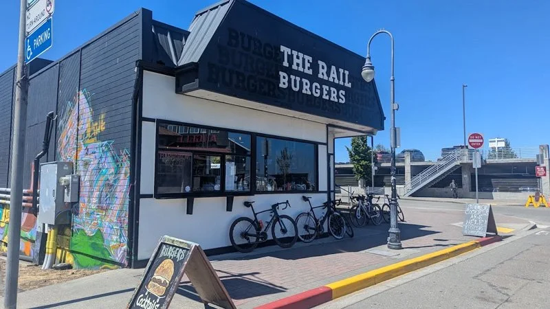 Small burger restaurant named 'The Rail Burgers' with bikes parked outside, a sidewalk, and a colorful mural on the side wall. Clear blue sky overhead.