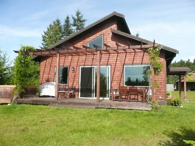 Red wooden house with a deck, sliding glass door, outdoor furniture, and surrounded by green grass and trees.