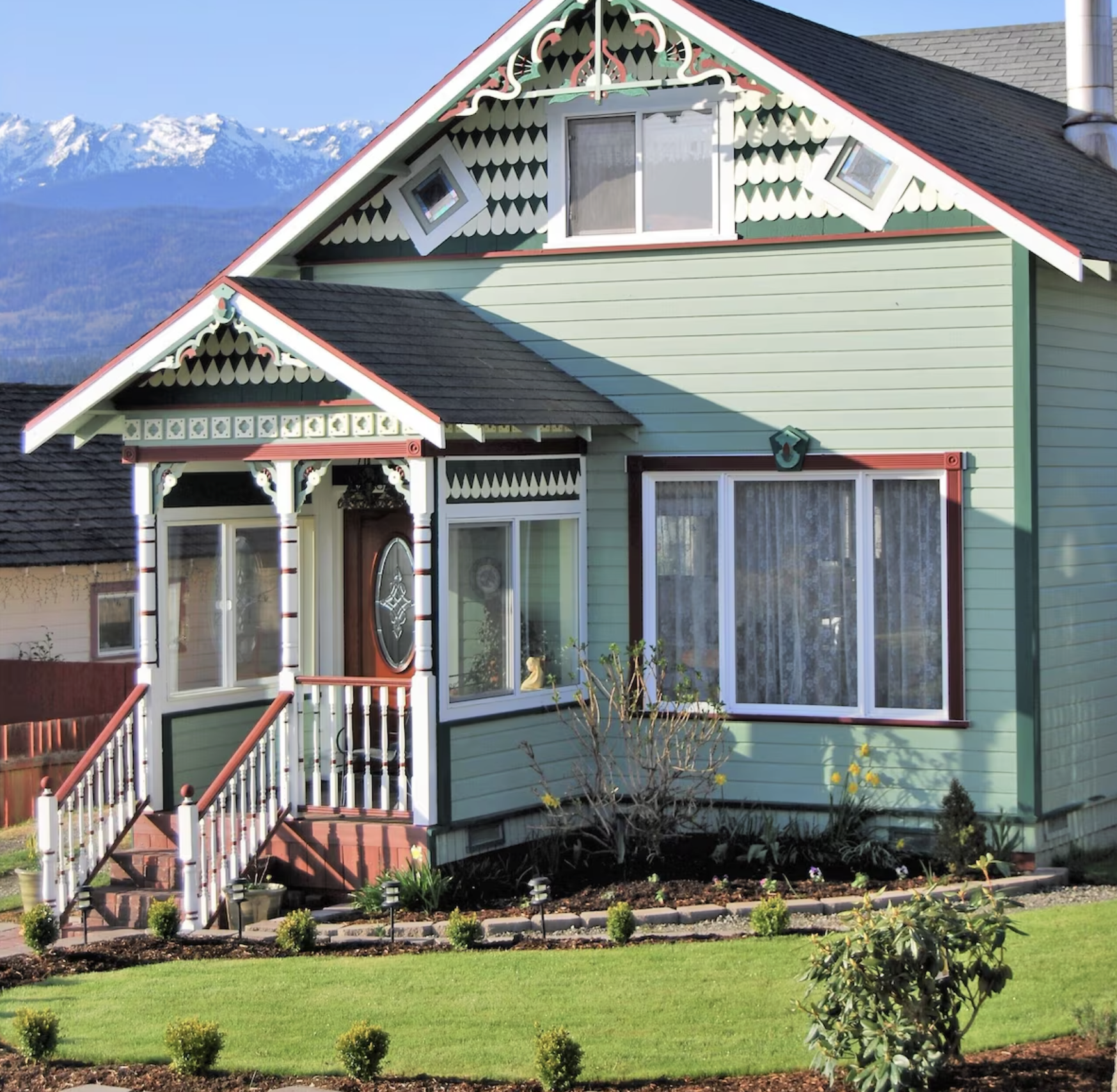 A two-story house painted in pastel green with decorative trim, front porch with stairs, large windows, and a well-maintained front yard with small bushes and flowers. Snow-capped mountains are visible in the background.