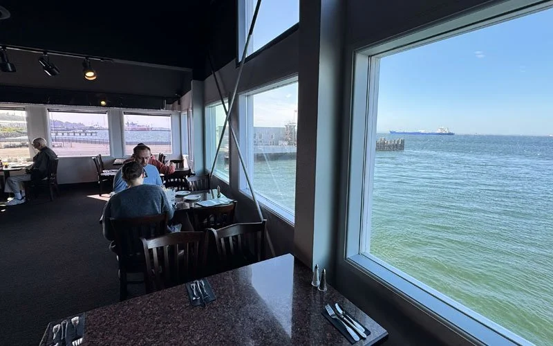 Inside a restaurant or cafe overlooking the water, with large windows showing a view of the ocean, ships, and a clear sky, and a few people dining.