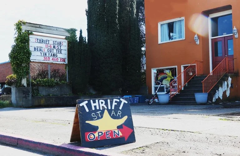 A sidewalk sign advertising Thrift Star is open, with the store name, a starfish graphic, and a red arrow pointing to the right, in front of a beige building with stairs and a bicycle.