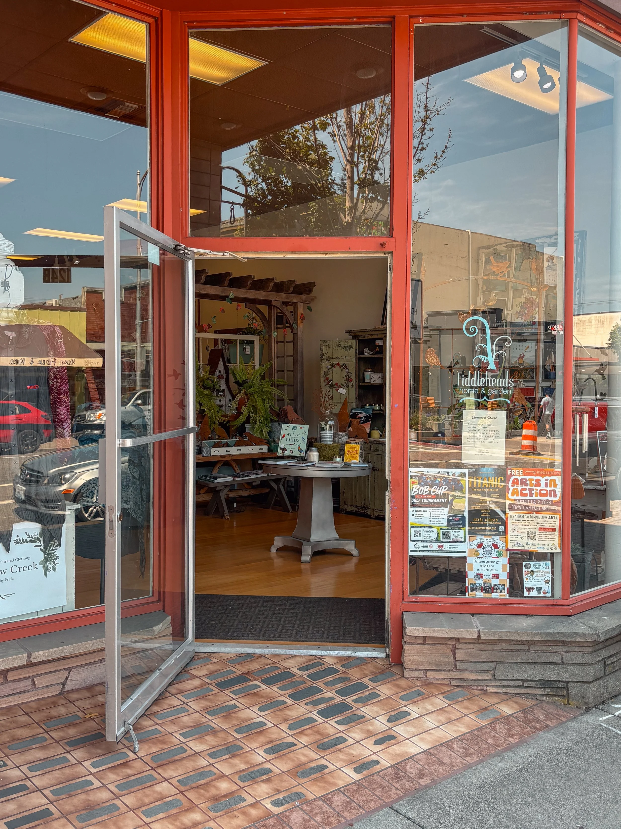 Exterior of a small shopping storefront named Fiddleheads with a sign indicating it is open, a flower box, and garden decorations visible inside.
