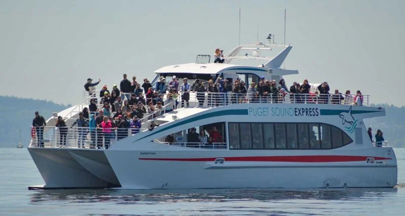 Large white ferry boat with many passengers on deck, labeled 'Puget Sound Express', on calm water under a cloudy sky.