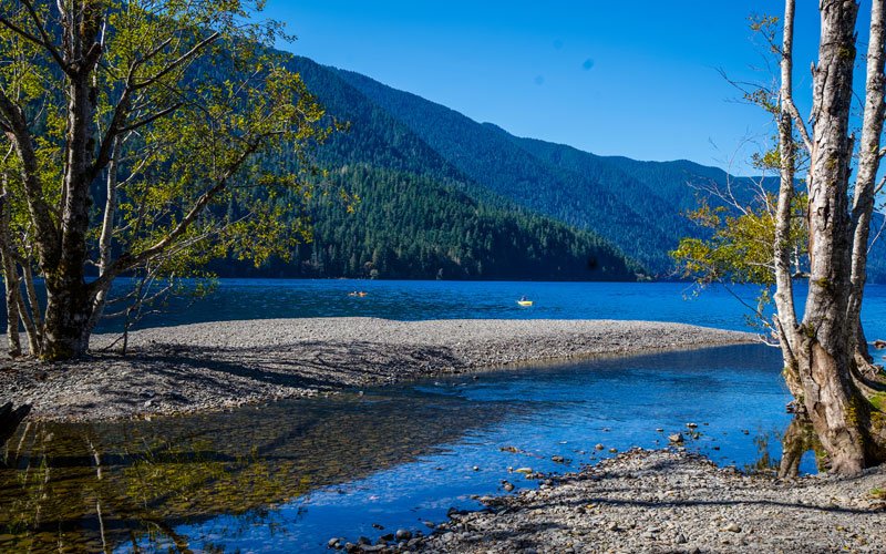 A peaceful lake scene with a pebble shoreline, trees, and distant mountains under a clear blue sky.