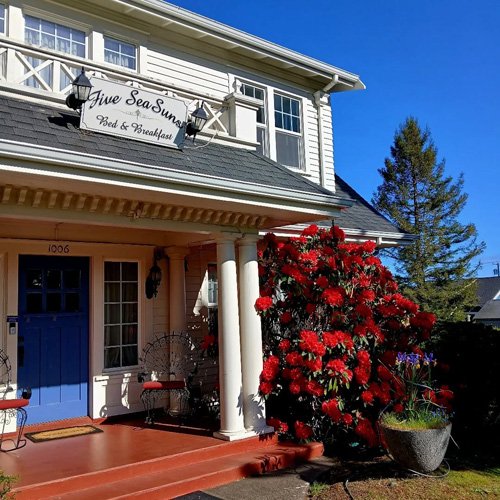 A two-story white building with a sign that reads 'Five Sea Suns Bed & Breakfast.' The front porch has a blue door, white columns, and outdoor seating. There is a large red flowering bush and potted plants near the entrance. The sky is clear and blue, and a large tree is visible in the background.
