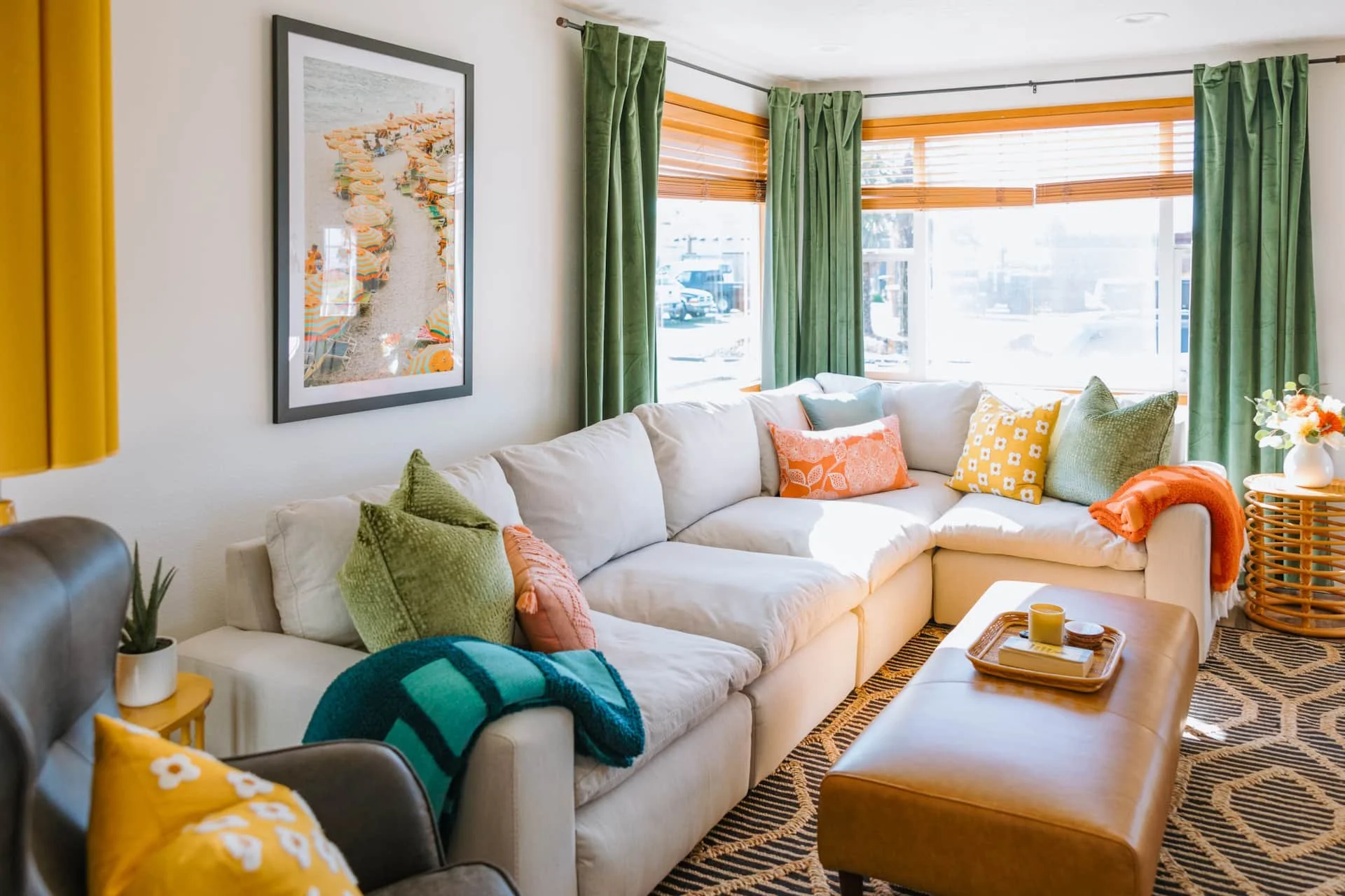 Living room with a large white sectional sofa decorated with colorful pillows, a wooden coffee table, and large windows with green curtains and wooden blinds, in a bright, sunny space.