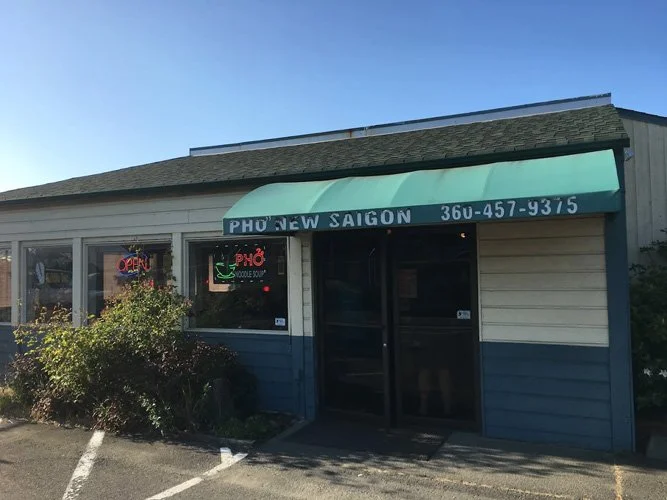 Exterior of a small Vietnamese restaurant called Pho New Saigon with a green awning, neon signs, and a parking space in front.