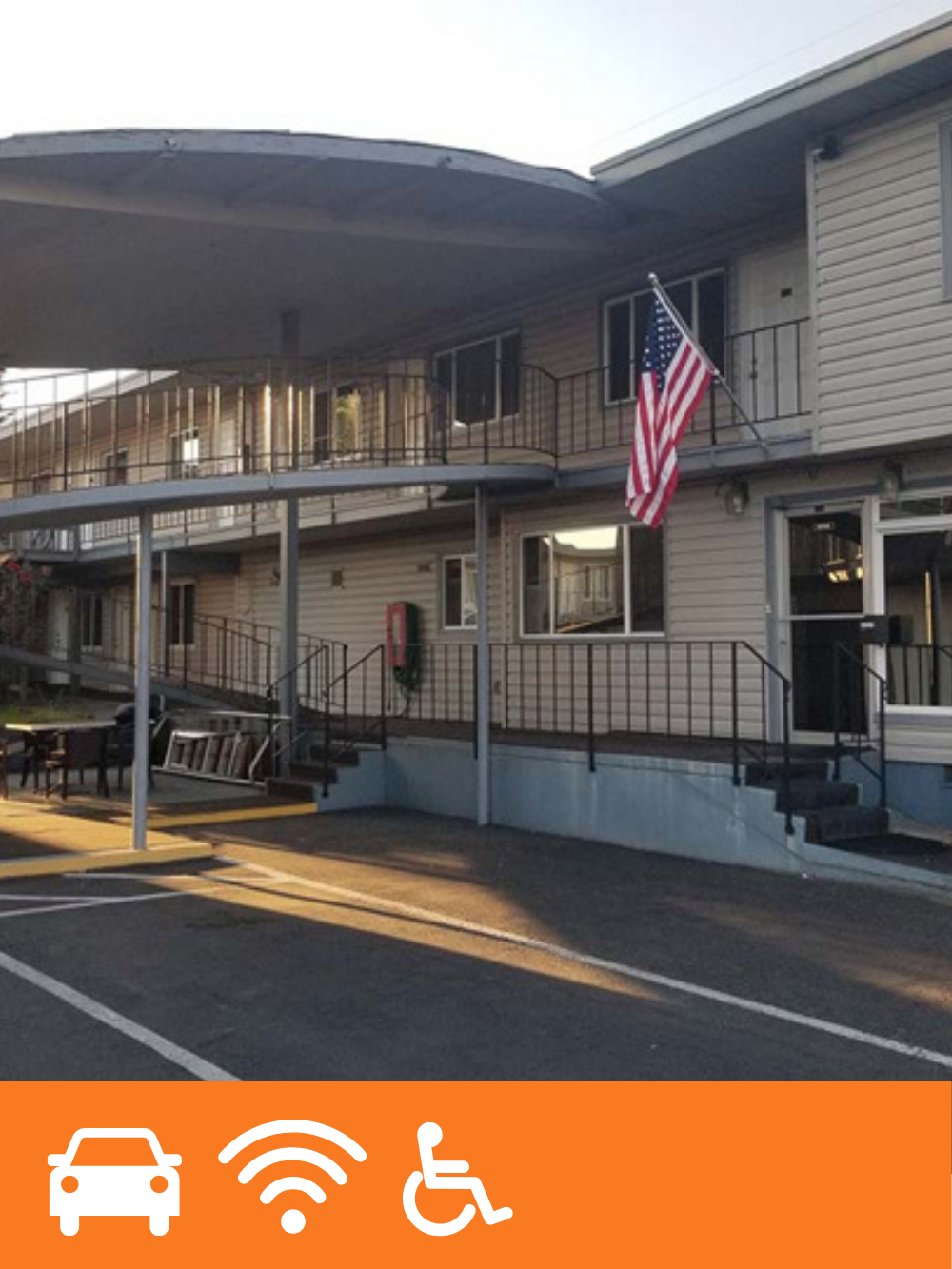 Two-story apartment building with beige siding, black railings, and a U.S. flag on a pole outside. There is an American flag hanging on the balcony, a fire extinguisher near the entrance, and a parking lot in front.