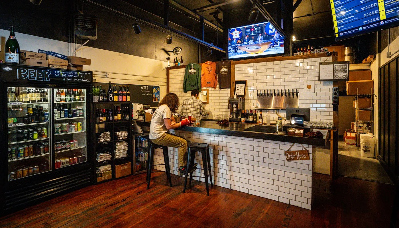 Interior of a bar or brewery with a person sitting on a barstool at the counter, bottles on shelves, and a bartender working behind the counter, with televisions displaying sports scores above.
