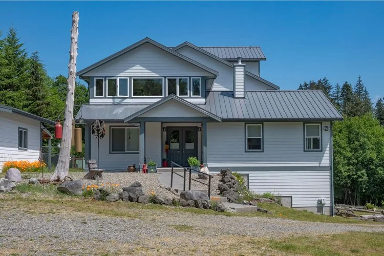 A modern two-story house with white siding, a covered front porch, and a metal roof, set against a backdrop of trees and a clear blue sky.