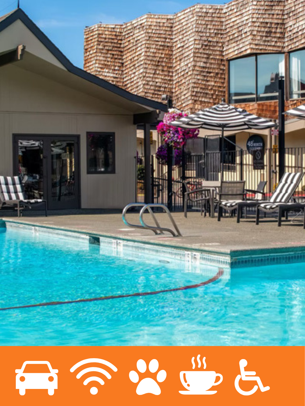 A swimming pool area next to a building with a sloped roof and wooden shingles. There are striped lounge chairs, a table with chairs, and umbrellas, with pink flowers hanging from a pole. The sky is blue with some clouds.