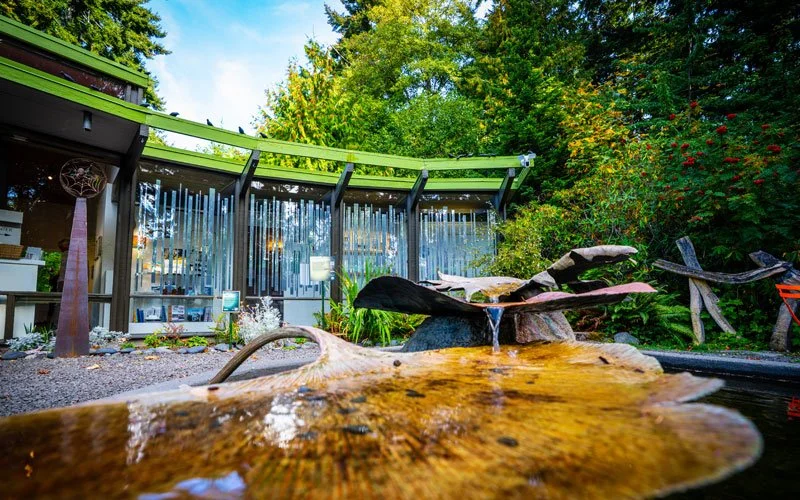 Outdoor scene with a modern building, a water fountain made of stacked flat rocks, surrounding greenery, and a glass wall with vertical decorative elements.