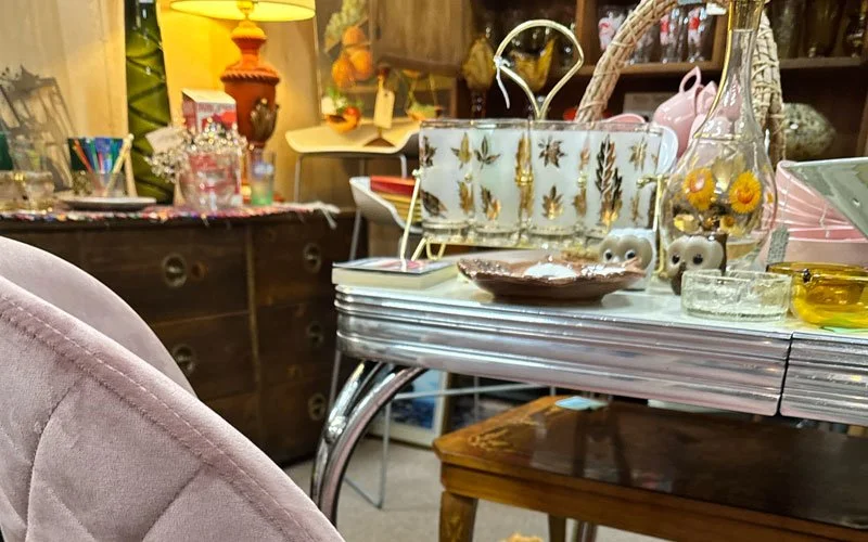A cluttered table filled with various decorative items and trinkets, including a white floral purse, small figurines, a dish, and glassware, in what appears to be a thrift store or antique shop.