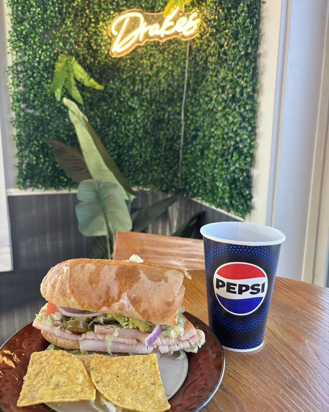 Inside of Drake's Pizza & Subs restaurant in Port Angeles with a neon sign, green wall decor, and a fast food meal in the foreground that includes a soda, sandwich, chips, and dip.