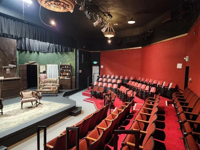 An empty theater with red upholstered seats arranged in rows, a small stage on the left with vintage furniture including armchairs and a bookshelf, green curtain backdrop, black walls with a red accent wall, and stage lighting overhead.