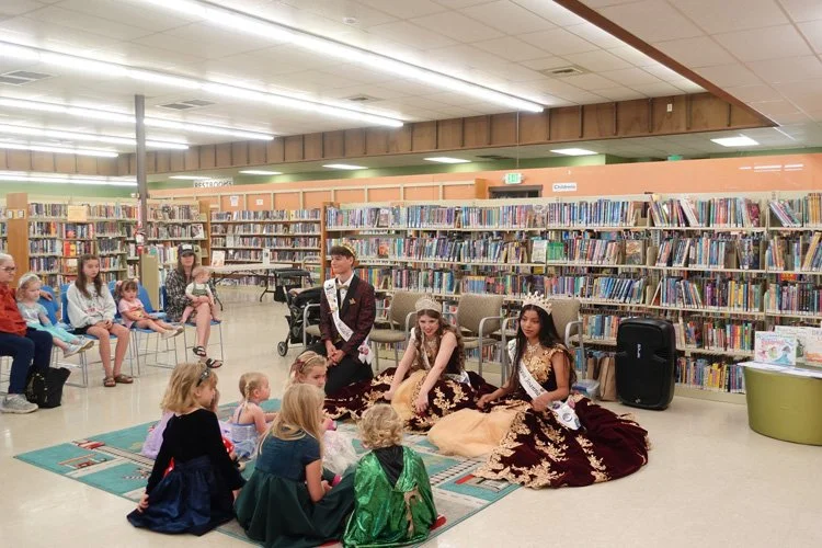 Children dressed as princesses and a young boy are participating in a storytime or presentation in a library, with shelves of books in the background and an audience of children and adults watching.
