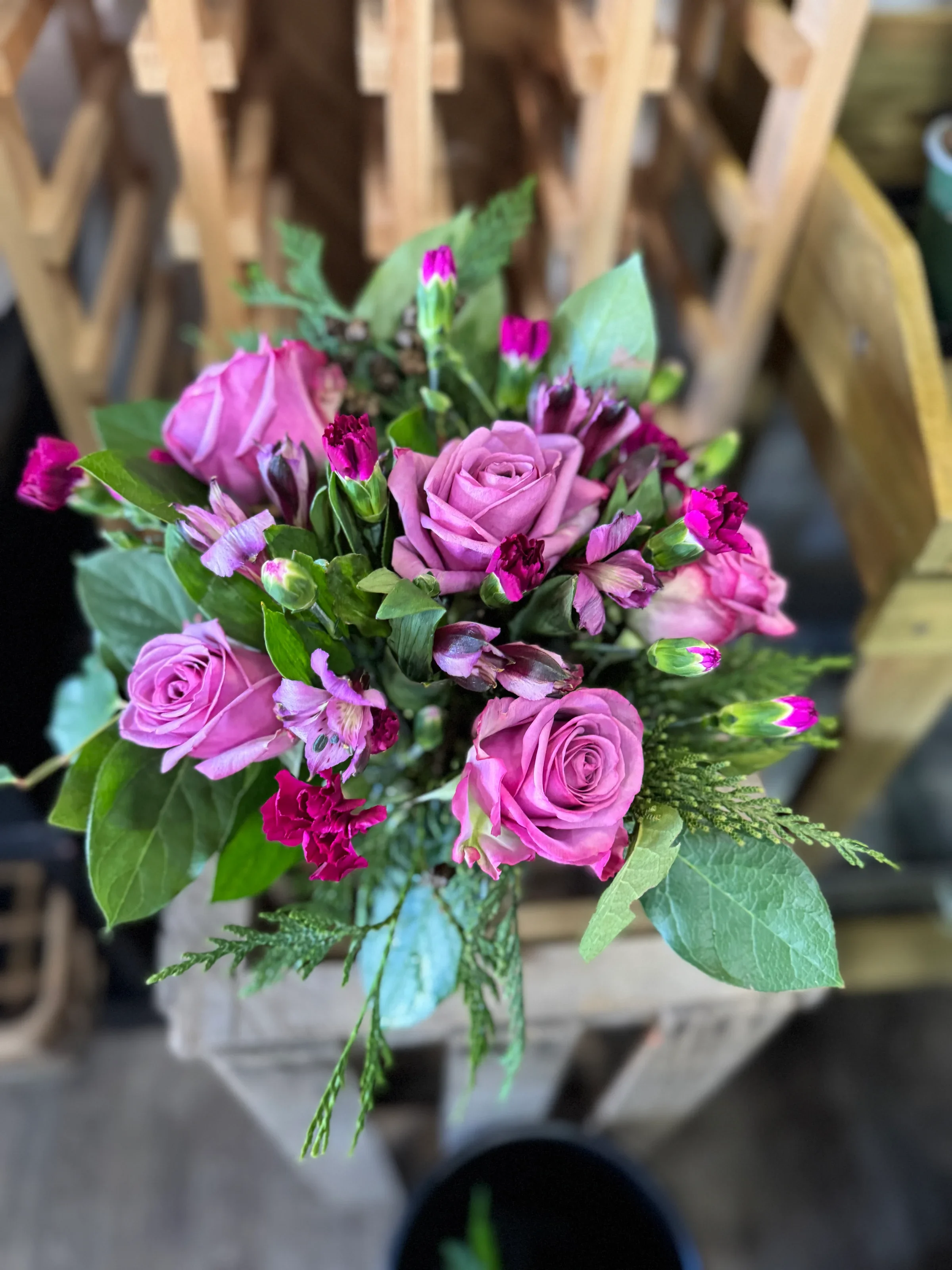 A large bouquet of pink, purple, and white flowers, including roses and carnations, in a white paper wrapper on a table near a window.