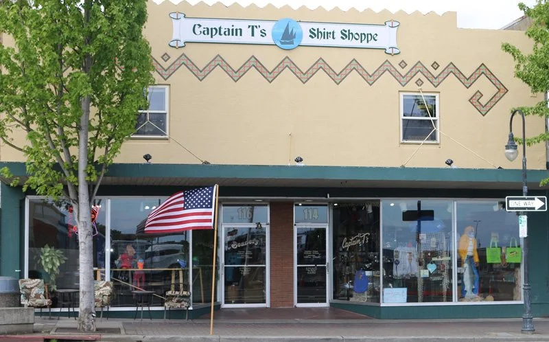 Exterior of Captain T's Shirt Shoppe with an American flag in front, large glass windows displaying clothing, and a sign with a sailboat logo.