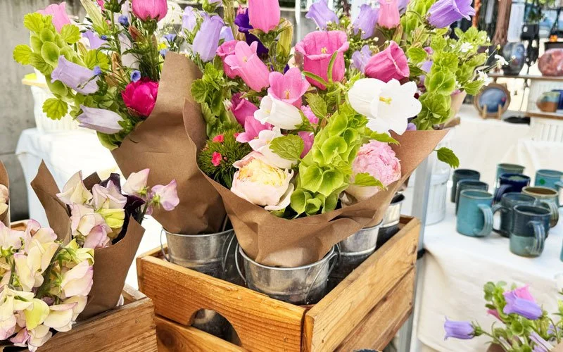 Bouquets of mixed flowers wrapped in brown paper with pink, purple, white, and green flowers in metal buckets on a wooden display at a market.