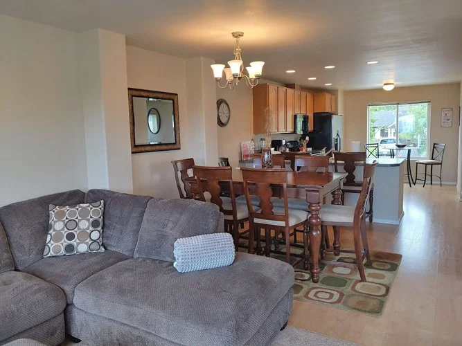 Open-concept living and dining area with a gray sectional sofa, a patterned throw pillow, a wooden dining table with chairs, a chandelier, a kitchen with wooden cabinets, black appliances, and a window with a view of the outdoors.