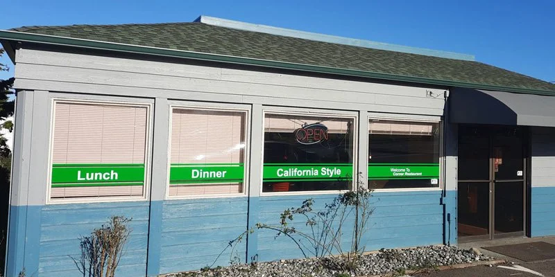 Exterior of a restaurant with a blue and white facade, large windows with green signs indicating lunch, dinner, and California style, a neon 'Open' sign in one window, and a glass door entrance with a dark awning.
