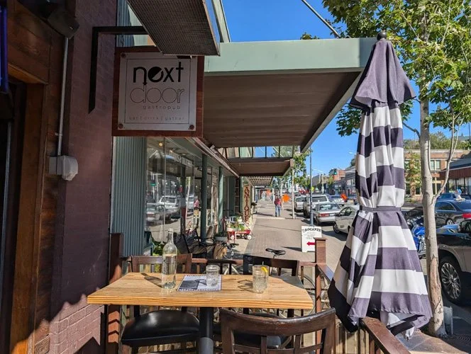 Outdoor patio of a cafe with a wooden table, chairs, and a large striped umbrella, in a small town street scene.