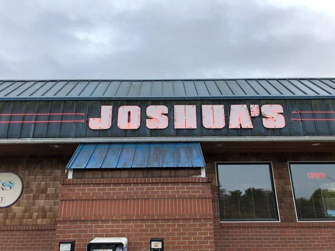 Sign on a brick building reads 'JOSHUA'S' in large, worn, pink letters with a metal roof and a small blue awning.