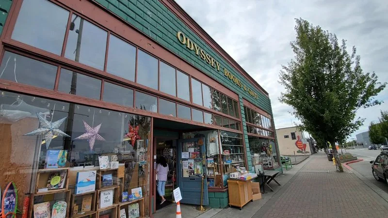 Exterior of Odyssey Book Shop with large glass windows displaying books and star-shaped decorations, and a tree-lined sidewalk outside.