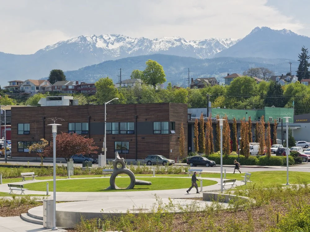 Urban park with sculpture, benches, lampposts, and people walking in front of a building, with green trees and snow-capped mountains in the background.