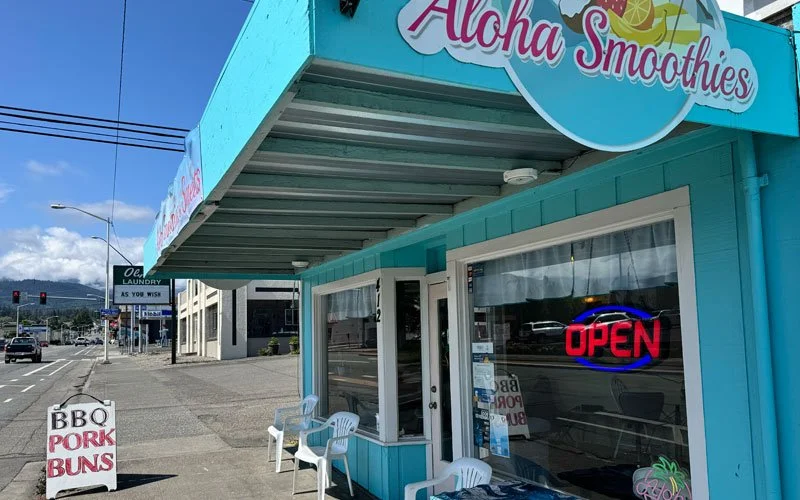 Aqua-colored smoothie shop named Aloha Smoothies with an open sign in the window, outdoor seating with white plastic chairs, a sidewalk sign advertising BBQ, pork, and buns, across the street from a busy road with cars and streetlights, mountains in the background.